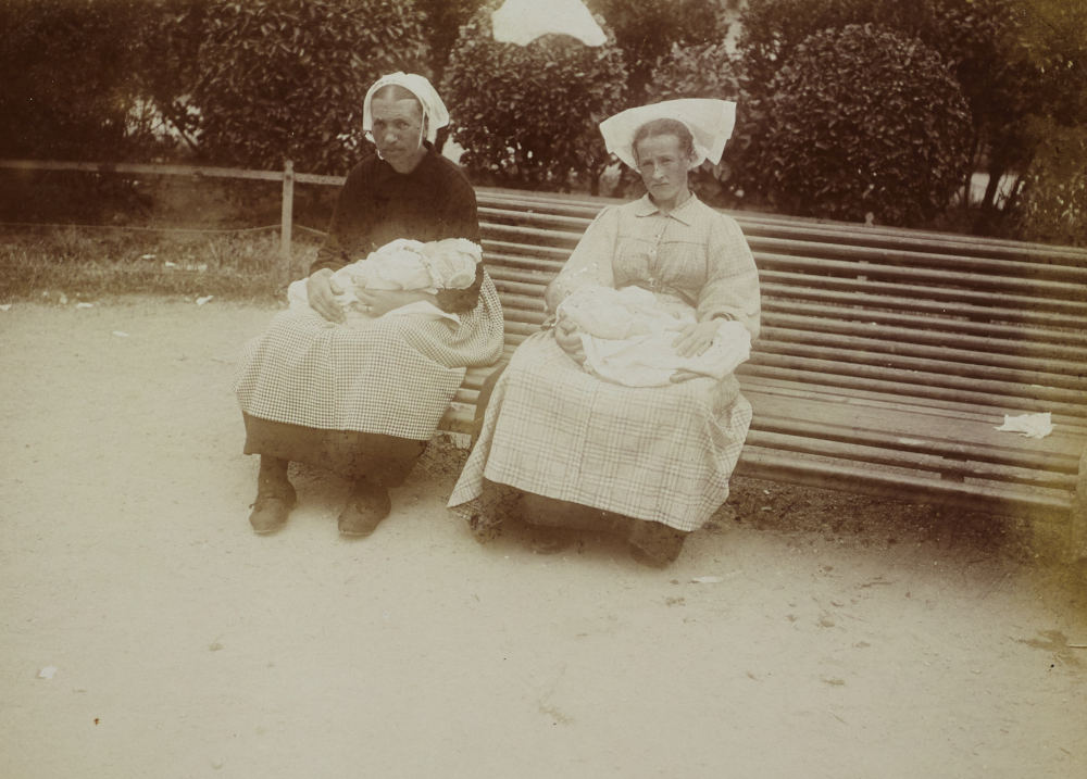 Deux nourrices assises sur un banc, chacune un bébé en lange sur ses genoux, dans le Morbihan. À gauche, la femme porte une coiffe de la presqu’île de Sarzeau, à droite, la jeune femme porte une coiffe de ville. Photo : Paul Géniaux. Source : collections du Musée de Bretagne, Rennes.