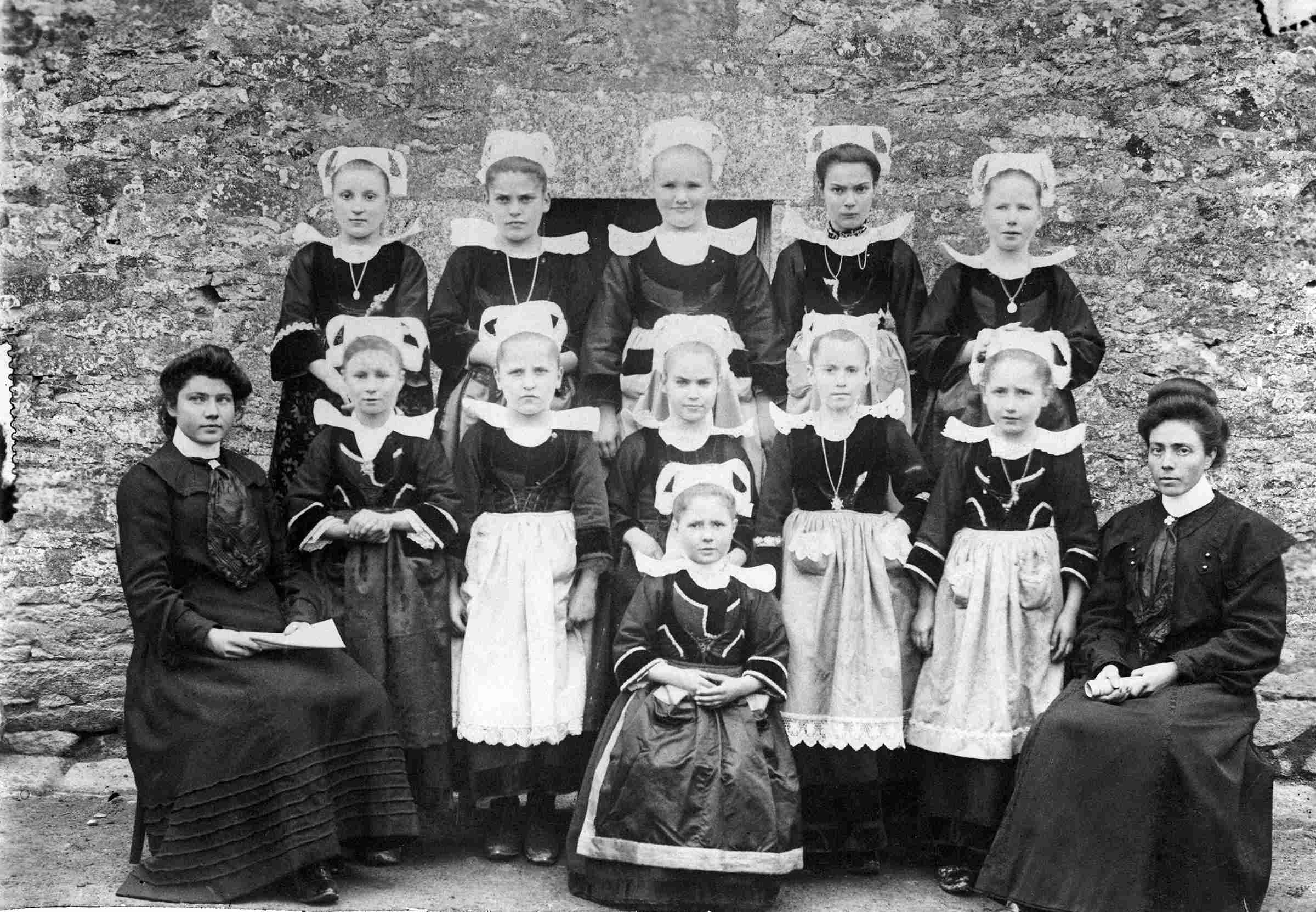 Une classe de filles à Kernevel (commune de Rosporden), vers 1910.  Les petites filles portent le costume traditionnel (Giz Fouen). Elles sont disposées sur deux rangs avec les deux institutrices, Mlle Sauban et son ajointe Eugénie Balcon, assises aux deux extrémités. Source : Collections du Musée de Bretagne.